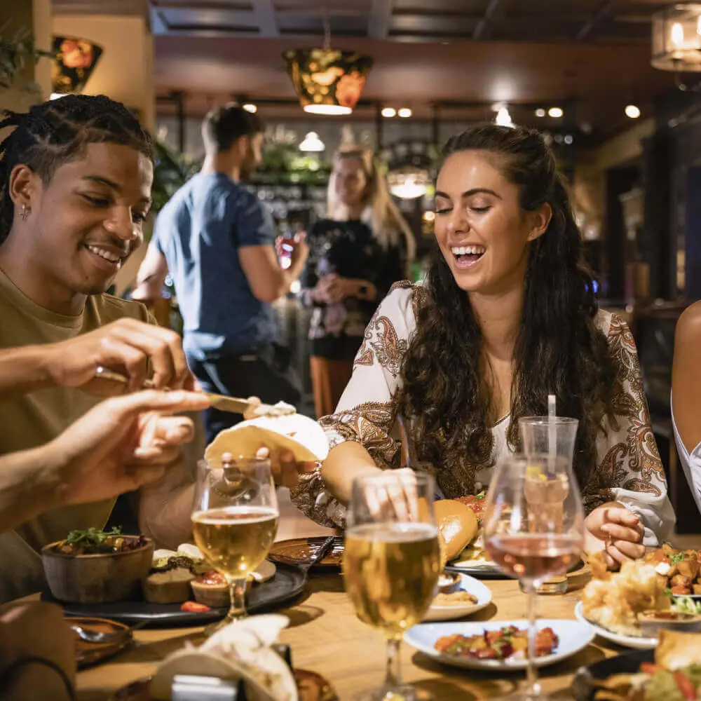 Young adults enjoy drinks and laughter at Village Clubs Camelback, sharing a meal in a cozy restaurant setting.