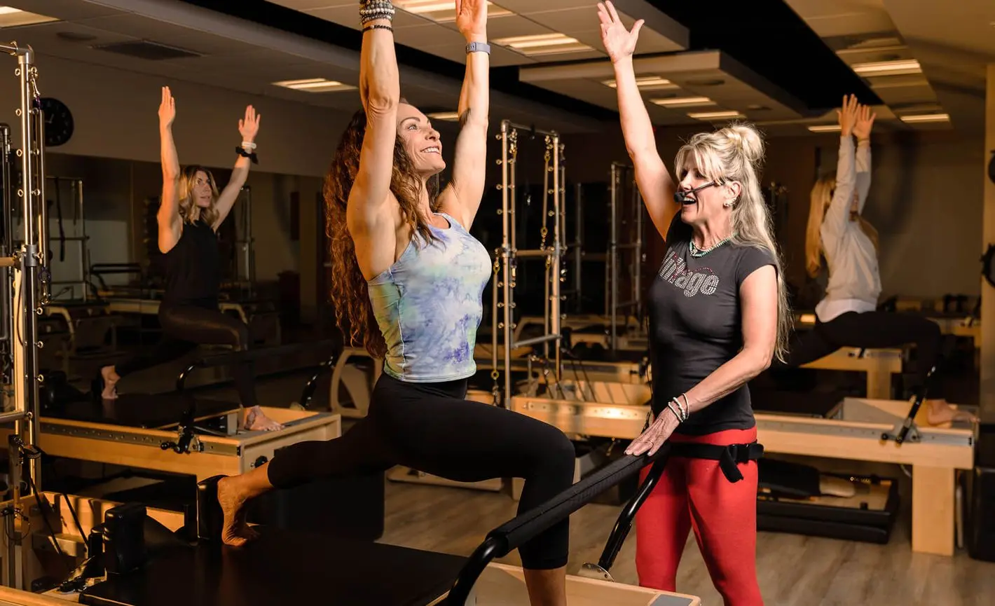 Village Clubs Camelback pilates class with instructor guiding a lunge pose on reformer, others practicing in bright studio.