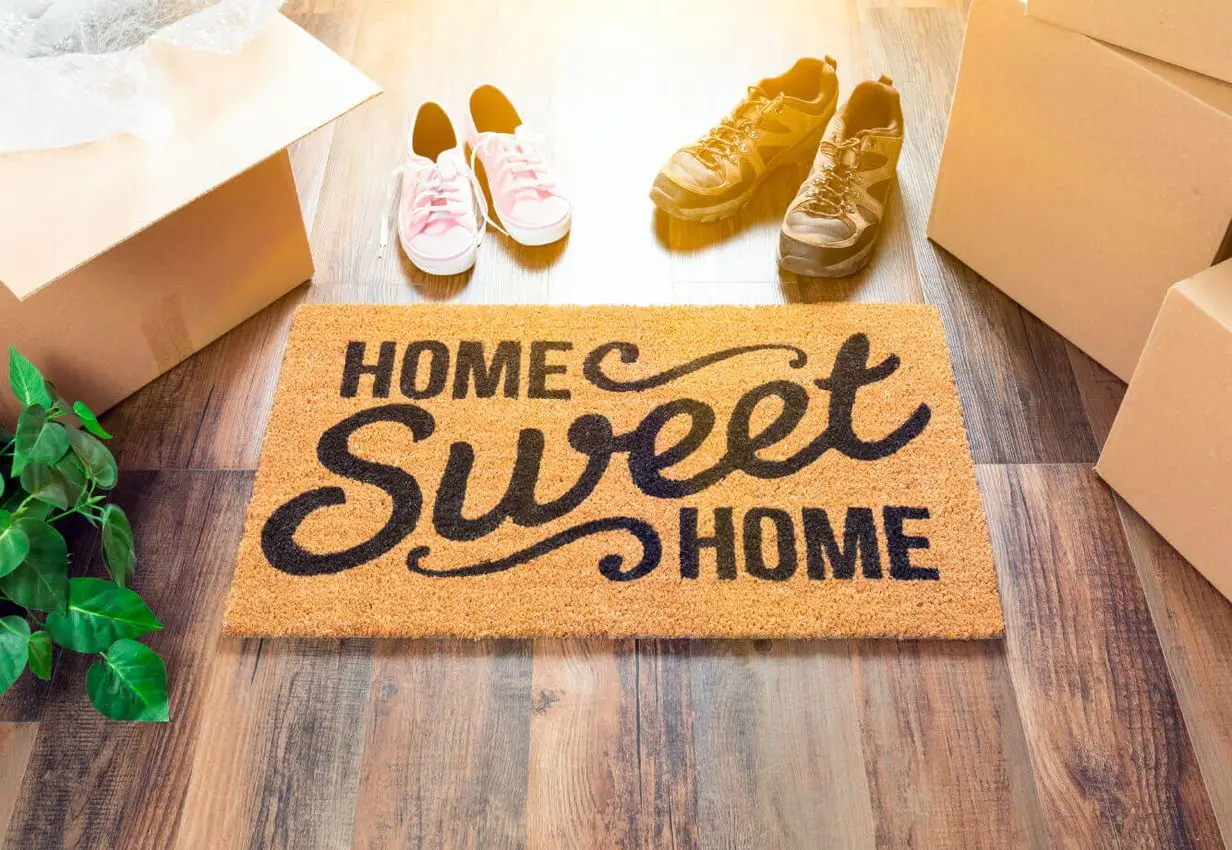 A welcoming Home Sweet Home doormat at Village Clubs Camelback, surrounded by boxes and shoes, highlights community support for moving.