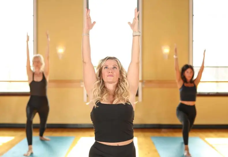 Three women practice yoga on blue mats at Village Clubs DC Ranch, in a bright studio with wooden floors and large windows.