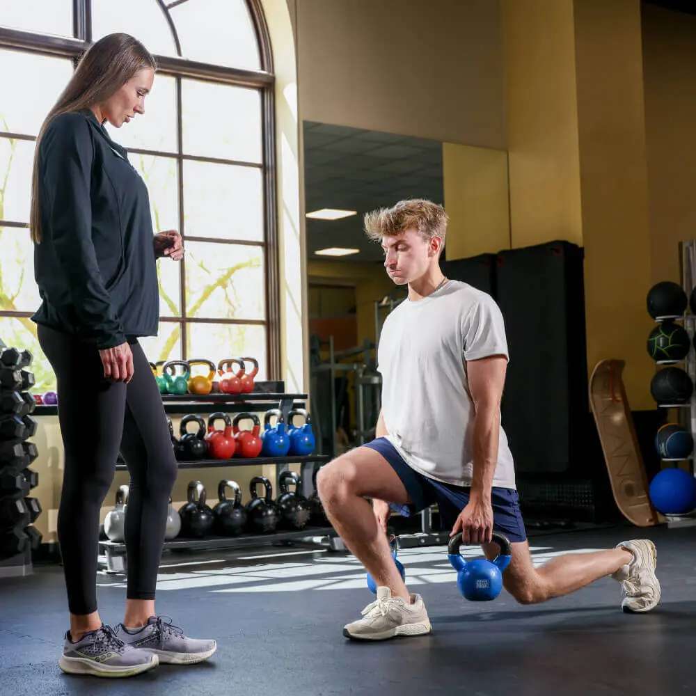 At Village Clubs Camelback, a man lunges with a kettlebell as a trainer observes, surrounded by gym equipment.