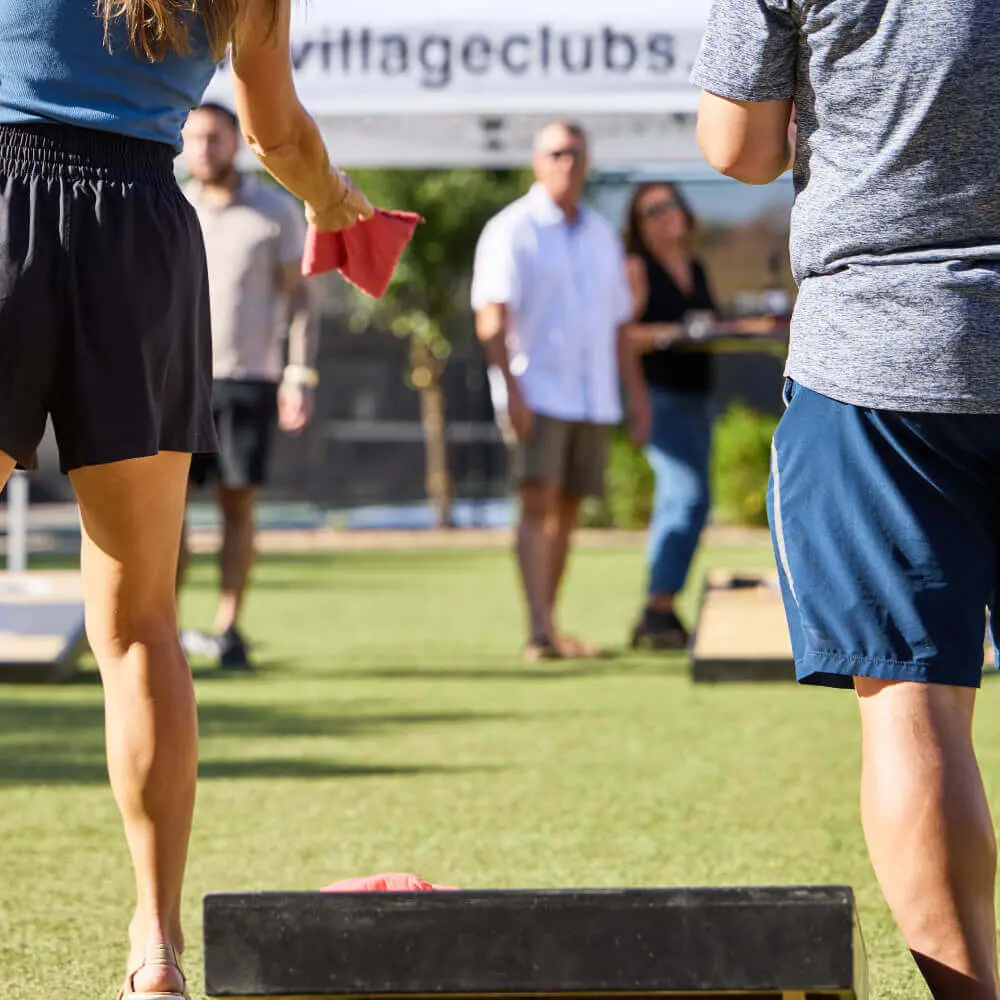 Members enjoy a sunny game of cornhole on the grass at Village Clubs Camelback in Arizona, with others socializing nearby.