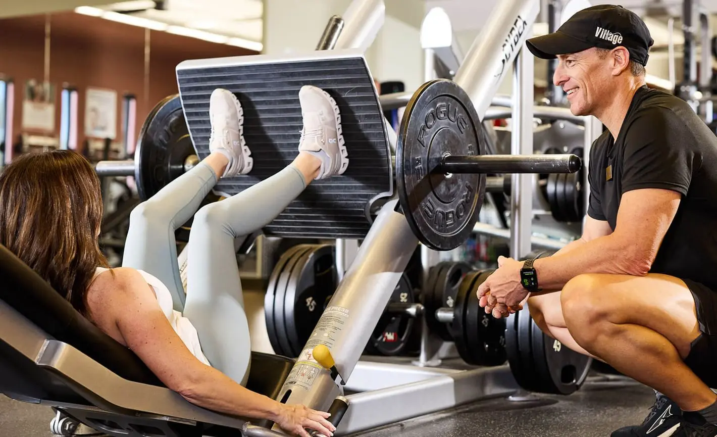 At Village Clubs Camelback, a woman uses the leg press as her trainer offers guidance; weight plates are loaded on the machine.