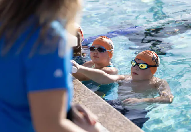 Two young swimmers in orange caps listen to an instructor at Village Clubs Camelback’s indoor pool in Arizona.