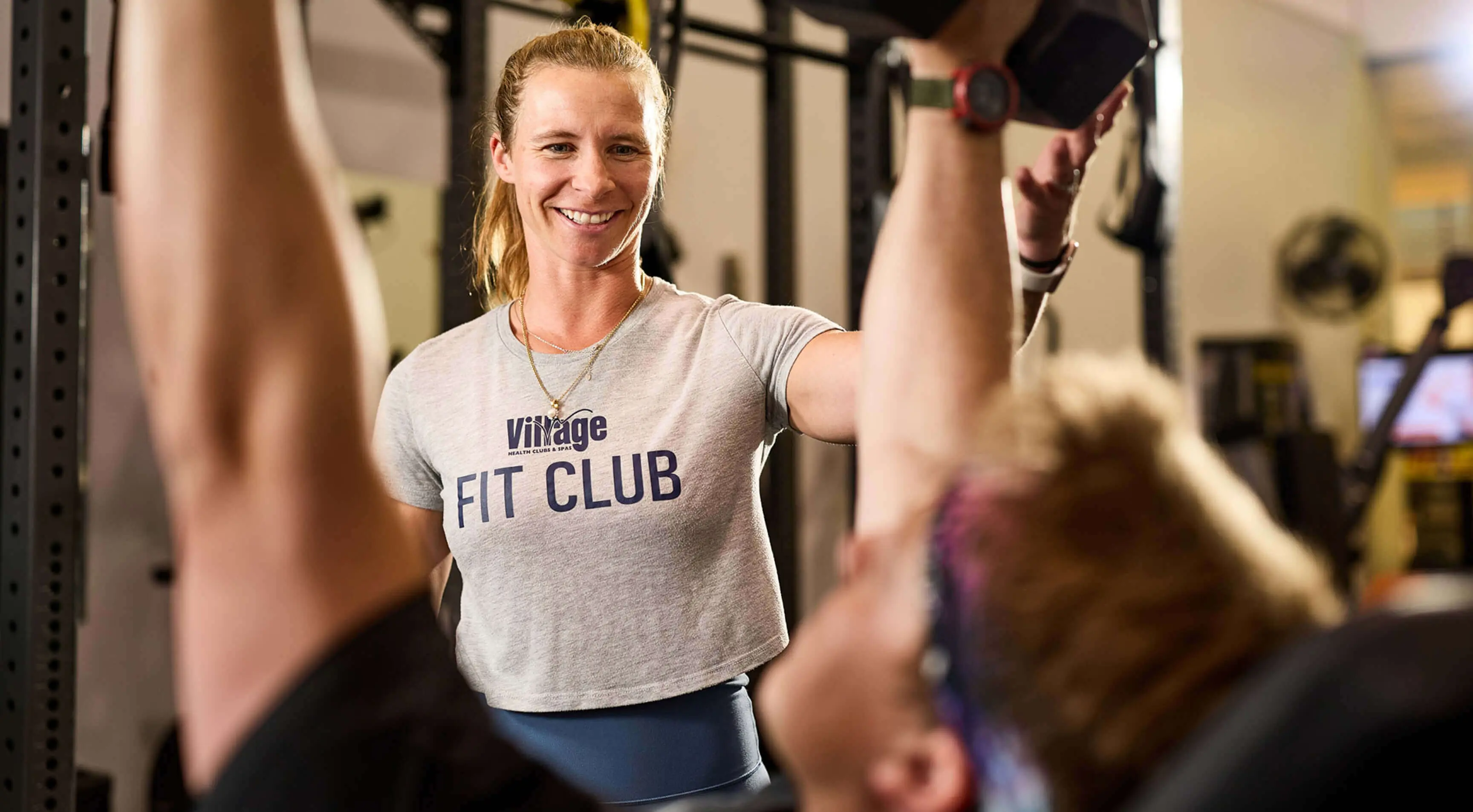 A Village Clubs trainer at DC Ranch, in a FIT CLUB shirt, supports and motivates a member lifting dumbbells in the gym.