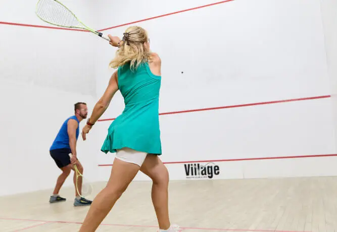 Two people play squash at Village Clubs Camelback. The woman swings her racket as the man watches, both in athletic gear on court.