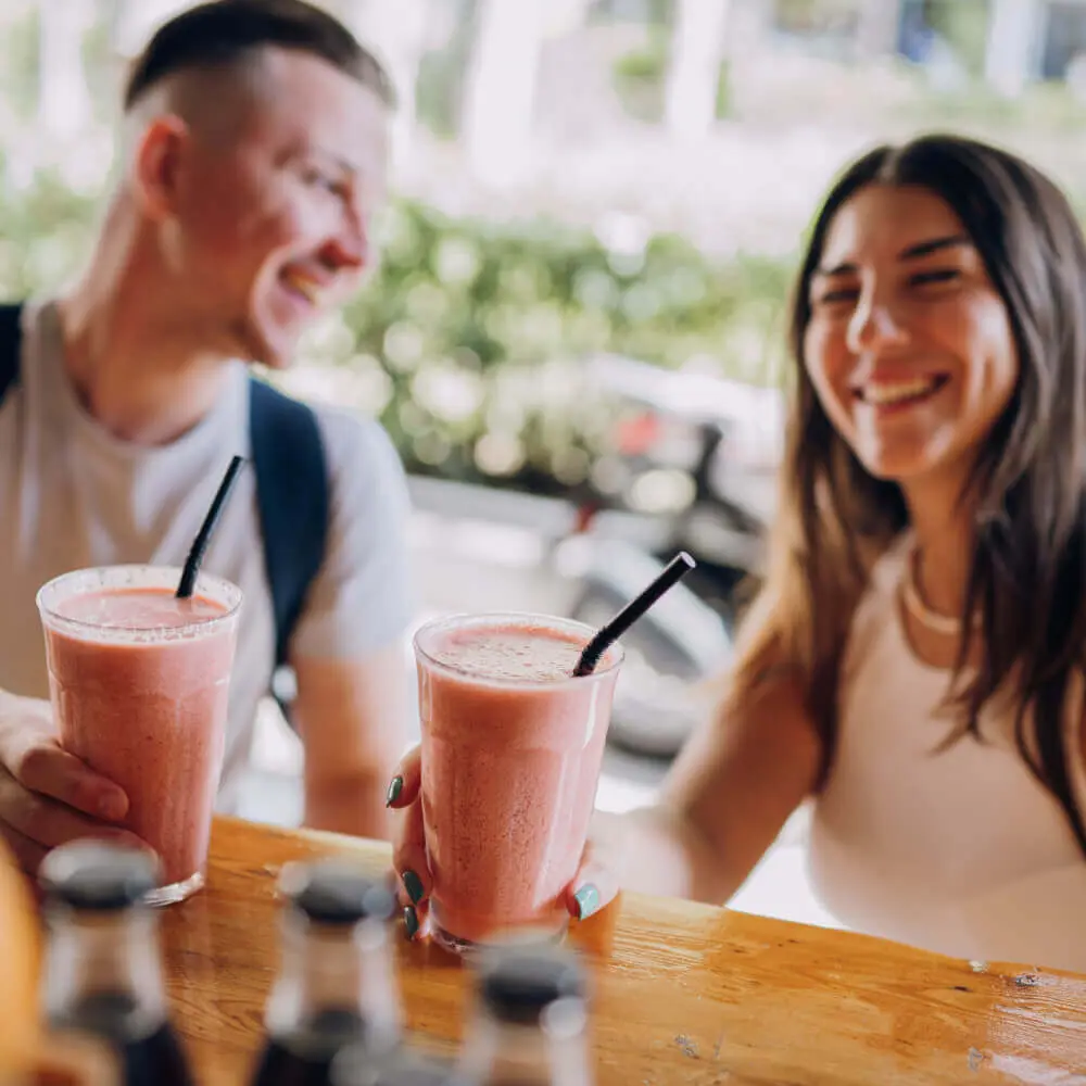 Guests at Village Clubs Camelback enjoy pink smoothies outdoors at a wooden table on a sunny day, bottles in the foreground.