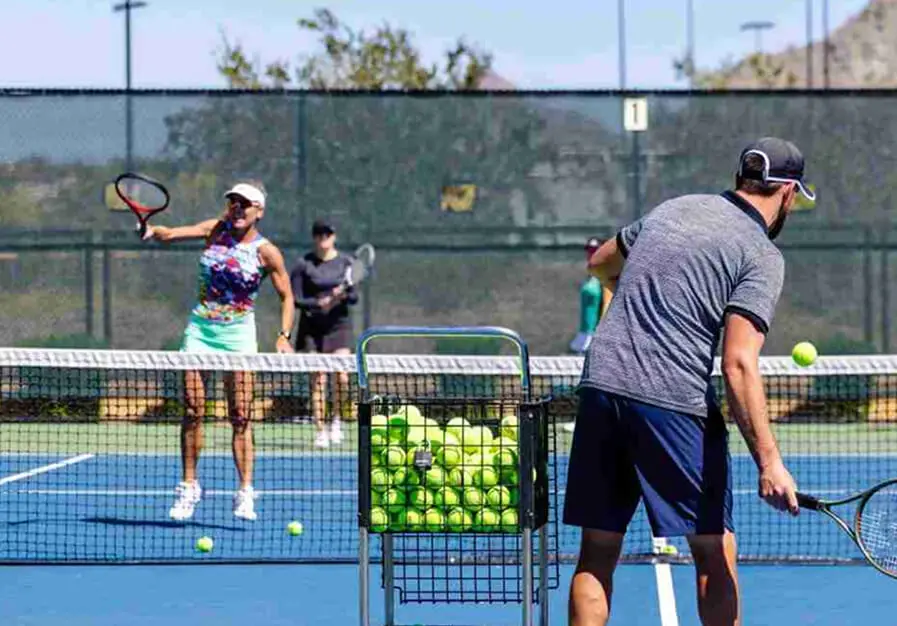Players enjoy a group tennis lesson at Village Clubs Camelback, with an instructor feeding balls on the outdoor court.