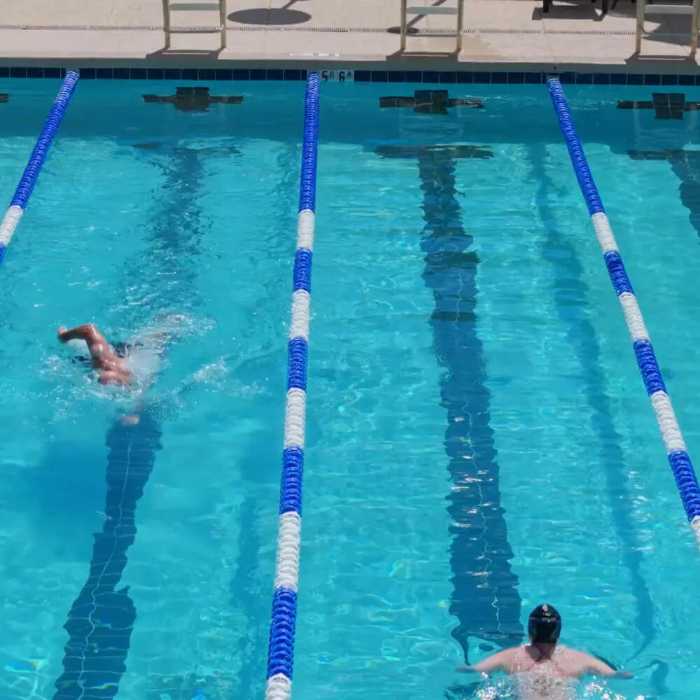 Two swimmers swim laps at Village Clubs Camelback’s outdoor pool, separated by blue and white lane dividers, with empty deck chairs nearby.