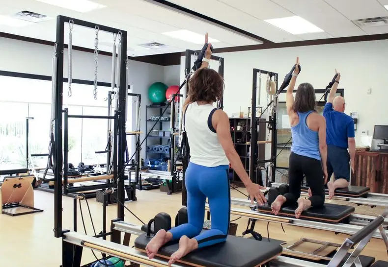 Members practice Pilates on reformers at Village Clubs Camelback, a bright studio with large windows, mirrors, and fitness equipment.