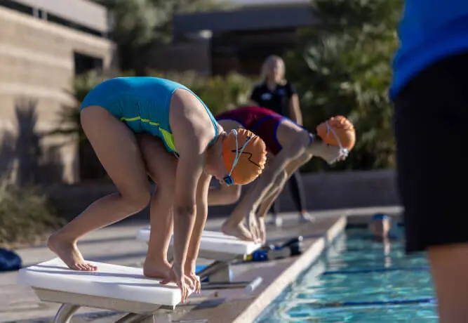 Swimmers at Village Clubs Camelback prepare to dive from starting blocks during a swim meet, coach watching by the pool.