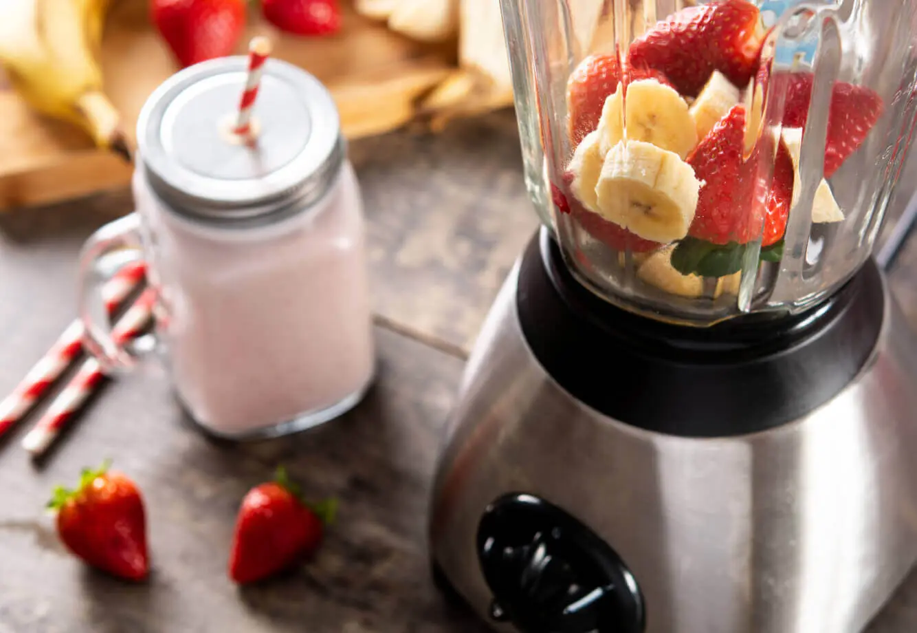 Village Clubs Camelback: Blender with bananas, strawberries, and a pink smoothie jar on a wooden table, fresh fruit in background.