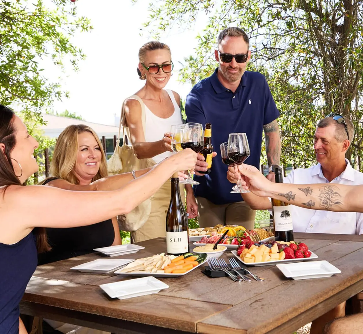 Members at Village Clubs Camelback enjoy a sunny outdoor toast with wine and snacks, gathered around a table under the trees.