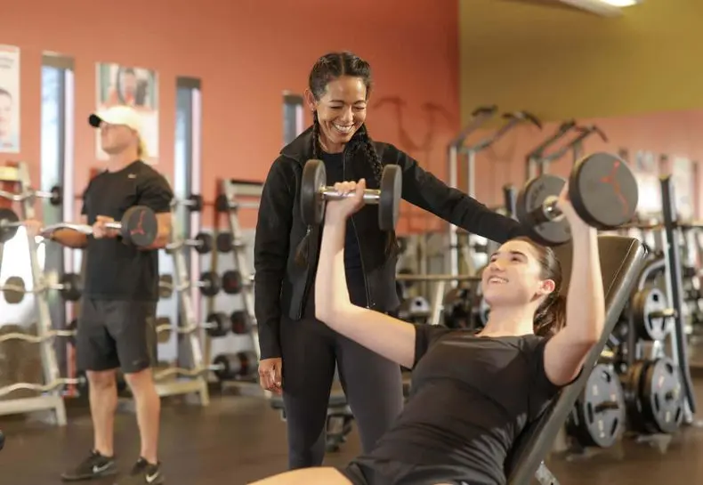 At Village Clubs Camelback, a trainer guides a woman lifting dumbbells on an incline bench in a well-equipped, warm-toned gym.