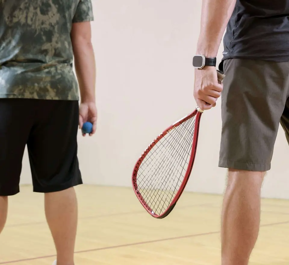 Two people prepare for a racquetball game at Village Clubs Camelback’s indoor court, holding a red racquet and blue ball.
