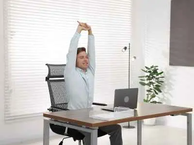 Young man in a light shirt sits at a desk with a laptop, stretching his arms overhead in a bright, modern office.