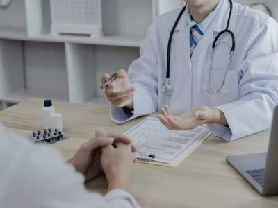 Doctor in a white coat with a stethoscope consults with a patient across a desk; documents on a clipboard are visible on the table.