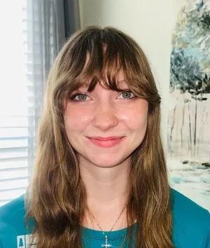 Young woman with long brown hair and bangs smiling at the camera, wearing a teal shirt and a cross necklace.