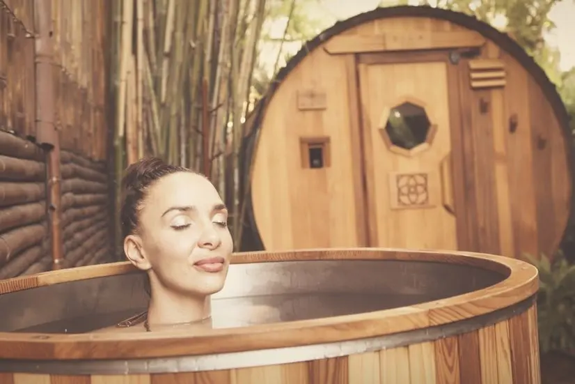 Woman relaxing in an outdoor cold plunge next to a wooden sauna barrel, part of a sauna and cold plunge experience in Miami, FL
