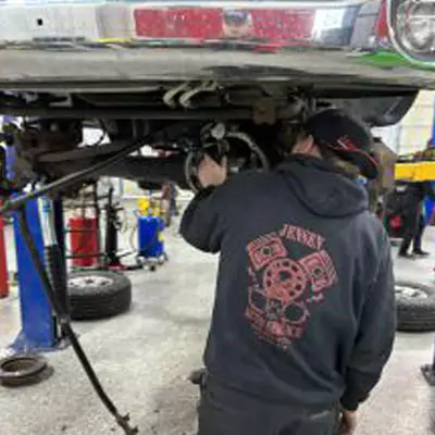 a man is engrossed in the task of repairing a car at an automobile repair shop. He is in a standing position, leaning towards the car while focusing on the intricate auto parts. The man seems to be a professional mechanic, given his diligent work on the vehicle. His attire is casual, with a noticeable logo on his shirt that hints at his affiliation with the repair shop. The car he's working on is parked outdoors, possibly on a ground near the shop.