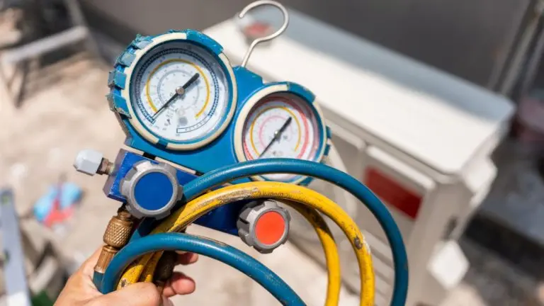 Technician's hand holding a manifold gauge with hoses in front of an outdoor air conditioning unit