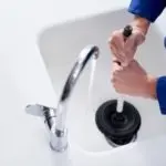 Person using a plunger on a kitchen sink, emphasizing regular drain cleaning by B&L Plumbing.
