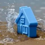 Blue toy house surrounded by splashing water and sand, illustrating plumbing issues related to flooding in Colorado Springs.