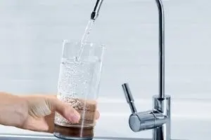 Hand holding a glass receiving filtered water from a modern kitchen faucet, illustrating plumbing solutions for home hydration.