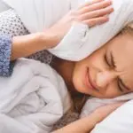 Young woman on a messy bed, hands covering ears, reflecting frustration with plumbing noise concerns. B&L Plumbing services.