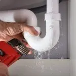 A person's hand using a pipe wrench to fix a leaking white PVC pipe, demonstrating plumbing maintenance by B&L Plumbing.