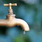 Brass faucet with dripping water, set against blurred green/blue foliage, highlighting plumbing services.