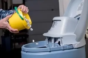 Person pouring salt pellets from a yellow bucket into a salt-based water softener, illustrating the maintenance process essential for effective hard water treatment.