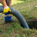 Worker using a vacuum hose to clear outdoor plumbing clogs in a grass-covered area, highlighting plumbing maintenance tasks.