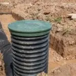 Cylindrical sewer main structure with green top in excavated dirt and gravel area during plumbing maintenance work.