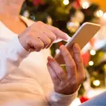 Individual in casual attire using a smartphone near a Christmas tree, representing B&L Plumbing’s holiday service reminders.