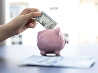 Hand inserting a hundred-dollar bill into a pink piggy bank, symbolizing savings and financial security.
