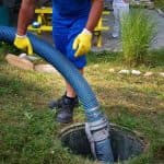 Worker in blue uniform conducting septic tank maintenance with vacuum hose in grassy area for B&L Plumbing services.