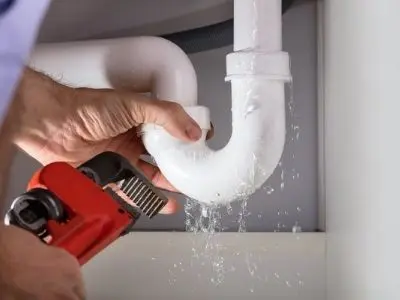 Person using a pipe wrench to fix a leaking white PVC pipe under a sink, illustrating evidence of plumbing problems and the need for timely repairs.