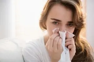 Woman holding a tissue to her nose, appearing distressed, in a bright indoor setting, illustrating common plumbing-related health concerns such as allergies or water quality issues.