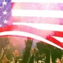 Crowd celebrating Fourth of July with raised hands in front of a waving American flag, symbolizing patriotism and community festivities.