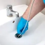 Person with blue glove using a flexible drain cleaning tool to clear a blocked sink for effective plumbing maintenance.