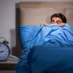 Young man in bed with blue bedding, looking fearful in a dim bedroom with a bedside clock, emphasizing plumbing concerns.