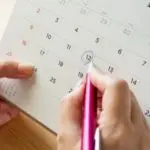 Hands writing important dates on a wall calendar with a pink pen, promoting plumbing maintenance habits by B&L Plumbing.