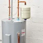 Water heater and expansion tank with copper piping against a textured concrete wall at Bud's Plumbing & Repair Service.