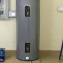Gray cylindrical water heater in a utility room with control panel, connected pipes, and a blue utility cart nearby.
