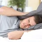 Man in bed covering head with pillow, showcasing stress amidst home improvement themes for Bud's Plumbing & Repair Service.