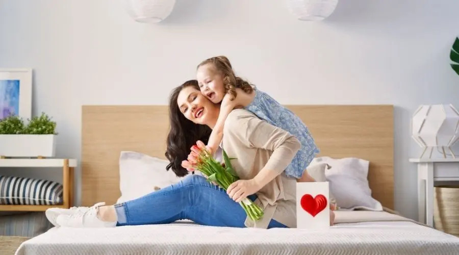 Mother and daughter celebrating Mother's Day, sitting on a bed with flowers and a heartfelt card, emphasizing home upgrades for thoughtful gifting.
