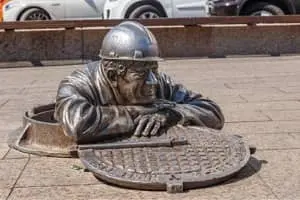 Bronze sculpture of a worker emerging from a manhole, representing outdoor plumbing maintenance and potential emergency plumbing issues.