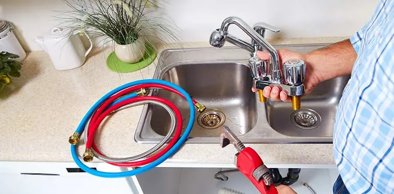 Plumber installing a faucet with red and blue supply hoses in a kitchen sink, emphasizing residential plumbing services by Lambert Plumbing in Port Moody, BC.