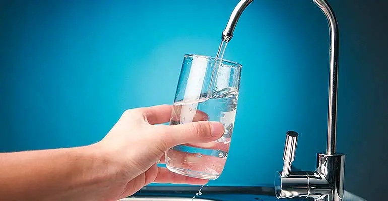 Hand holding a glass under a water faucet, demonstrating clean drinking water from a whole house filtration system.